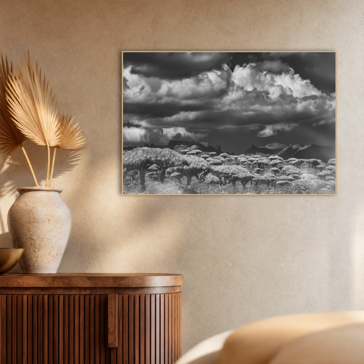 Gito.Art - Exclusive Art Print - Photography by Delyan Markov
A framed black and white landscape art print depicting a dramatic cloudy sky over a grassy field, displayed on a textured beige wall above a wooden sideboard with a vase of dried palm leaves.