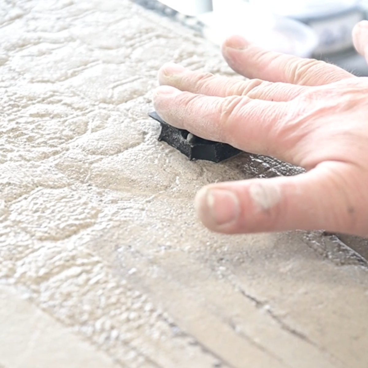 A close-up of a textured beige stone or concrete surface with a hand pressing a small black rectangular tool into it.