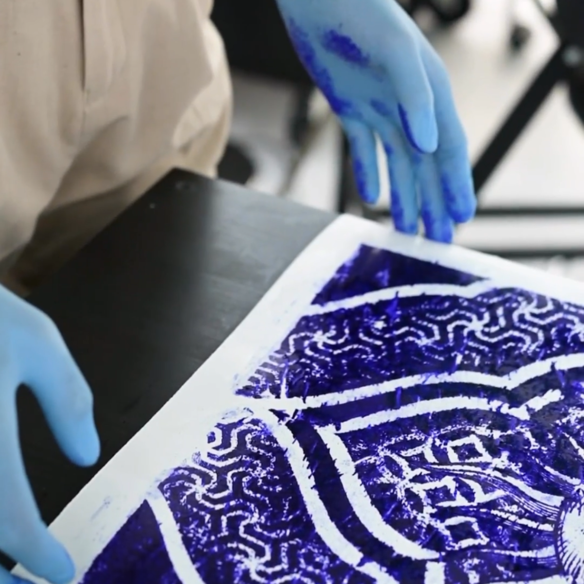 A close-up of a person wearing blue gloves handling a square sheet of artwork printed with intricate white patterns over a deep blue background, set on a dark table.