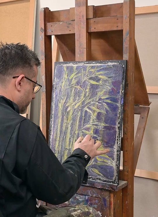 An artist in a black shirt and glasses is painting on a canvas with yellow and purple brushstrokes, mounted on a wooden easel in an art studio.