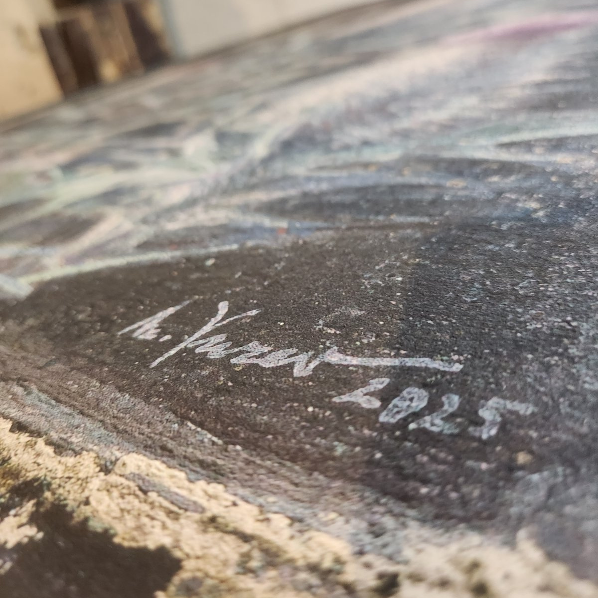 Close-up of a textured charcoal or blackboard surface with a white signature reading 'h. Yasser 2025' and a blurred painted scene in the background.