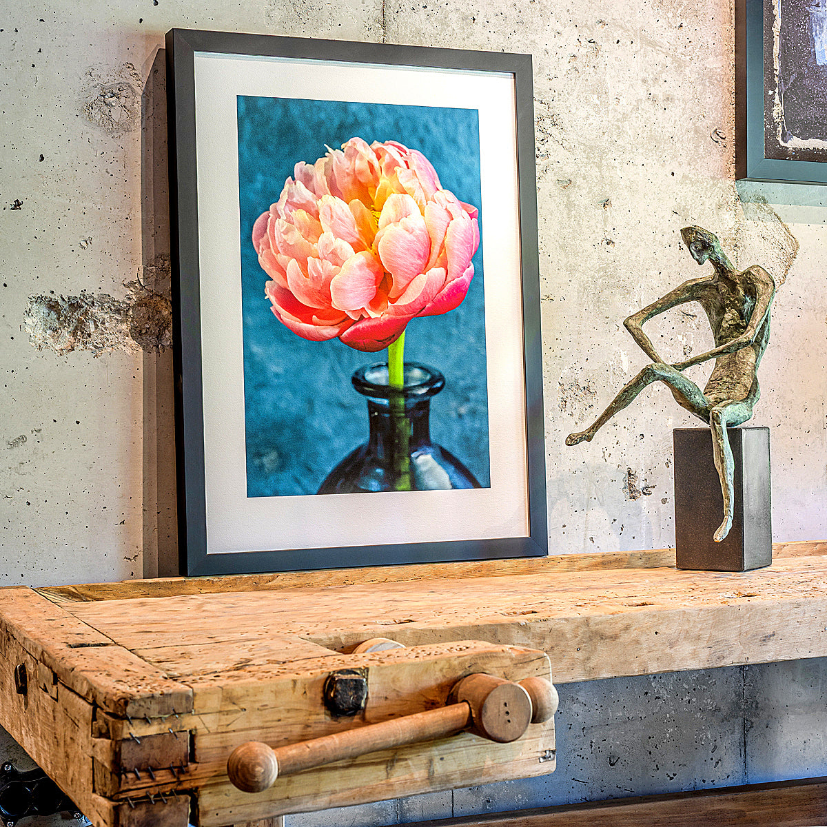 A framed art print featuring a pink peony flower in a dark glass bottle against a blue background, displayed on a rustic wooden table next to a bronze figurine sculpture in a styled interior setting.