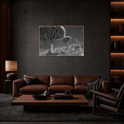 Gito.Art - Exclusive Art Print - Photography by Delyan Markov
A framed black and white landscape photograph of quiver trees in a desert, displayed on a dark textured wall above a brown leather sofa in a modern, dimly lit living room.